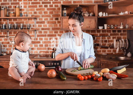 Happy young family cooking breakfast together. Mother chopping cucumber on cutting board while her son sitting near on kitchen table Stock Photo