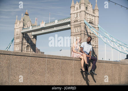 Young couple sitting on wall, smiling, Tower Bridge in background, London, England, UK Stock Photo