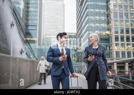 Businessman and businesswoman using mobile phone and pulling trolley luggage, Canary Wharf, London, UK Stock Photo