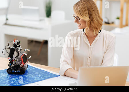 Feeling grateful for your help. Positive upbeat smart engineer working in the office and using laptop while collaborating with the automatic robot and Stock Photo
