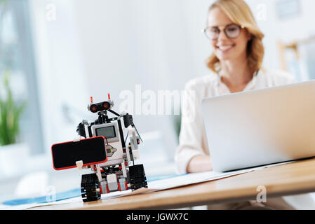 Mastering human skills. Futuristic automatic funny robot standing on the table in the office and holding digital gadget while engineer using laptop on Stock Photo