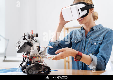 In the parallel world. Smiling happy joyful woman sitting in the lab and using visual reality glasses while enjoying electronic robot Stock Photo