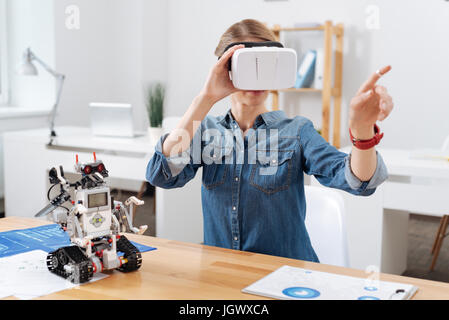 Full of interest. Concentrated confident skilled volunteer sitting in the lab and using visual reality glasses while interacting with electronic robot Stock Photo