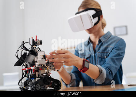 Touch the other reality. Curious young pleasant woman sitting in the lab and using visual reality glasses while interacting with automatic robot Stock Photo