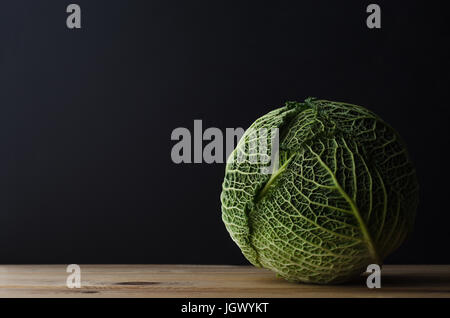 A whole head of leafy green cabbage on a wood plank table against a black chalkboard background. Moody lighting. Stock Photo