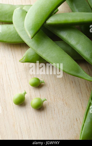 A pile of pea pods, with three tiny baby peas extracted and placed on a light wooden table.  Stalks intact.