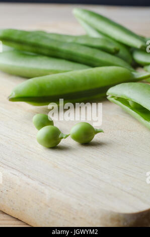 Close up of fresh, raw pea pods, scattered on a wooden chopping board, with three small peas extracted and placed in the foreground with stalks intact