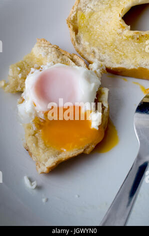 A partly eaten free range fried egg with yolk spilling onto bagel and plate below.  Fork visible.  Shot from diner's perspective. Stock Photo