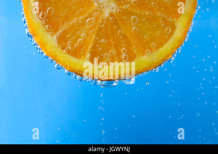 Close up of a fresh orange slice at top of image and facing forwards, suspended in sparkling bright blue water.  Oxygen bubbles float upwards and cove Stock Photo