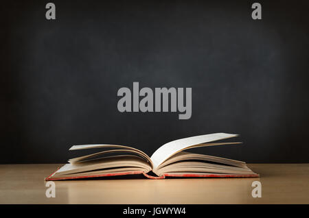 An old, worn, fabric covered red text book, lying opened on a school or college classroom desk.  Blackboard background provides copy space. Stock Photo