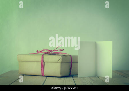 A gift box wrapped in brown paper and tied with pink raffia bow on a wooden table.  An opened, blank  greeting card faces front.  Cross processed to g Stock Photo
