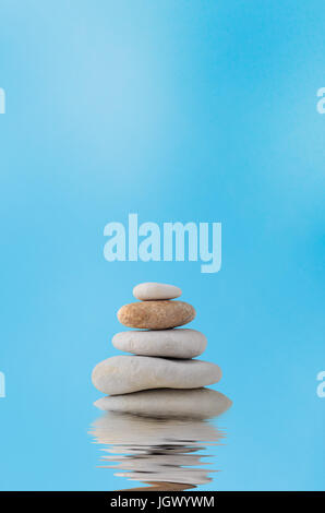 A pile of five stacked stones with a watery rippled reflection below and blue sky with light white cloud haze in the background. Stock Photo