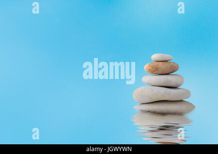 A pile of five stacked stones with a watery rippled reflection below and blue sky with light white cloud haze in the background. Stock Photo