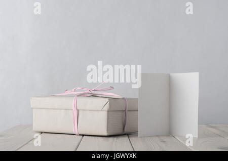 Photo of a gift box simply wrapped in brown paper, tied with pale icy pink raffia on an old planked wooden table. A blank open greeting card faces fro Stock Photo