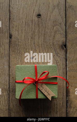 Overhead shot of a simple green gift box on an old oak wood planked table, tied to a bow with red satin ribbon, with a vintage style 'Merry Christmas' Stock Photo