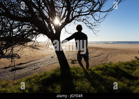A silhouetted man leans on a tree as the morning sun beams through the branches at a surf beach in Port Macquarie, Australia. Stock Photo
