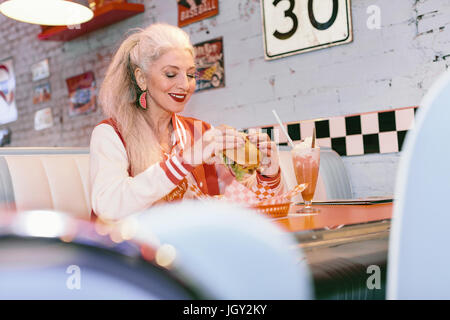 Mature woman in baseball jacket eating burger in 1950's diner Stock Photo