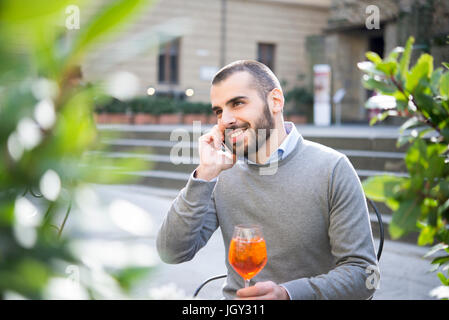 Young man sitting outside bar, holding drink, using smartphone Stock Photo