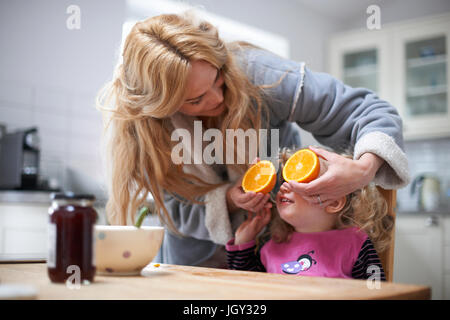 Young girl sitting at kitchen table, mother holding halved orange in front of daughter's eyes Stock Photo