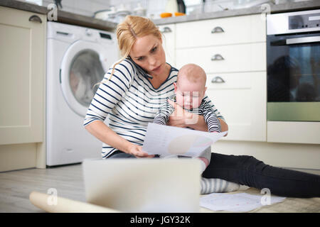 Woman holding baby son in arms, whilst looking through work on kitchen floor, speaking on smartphone and looking at laptop Stock Photo