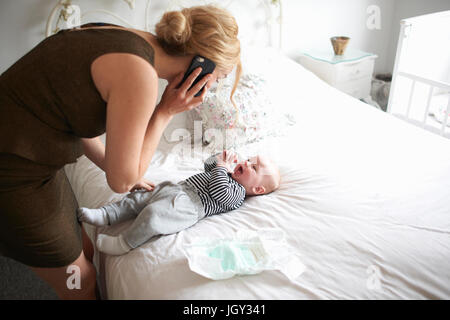 Mother changing baby son's nappy while using mobile phone Stock Photo