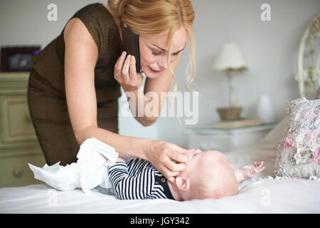 Mother changing baby son's nappy while using mobile phone Stock Photo