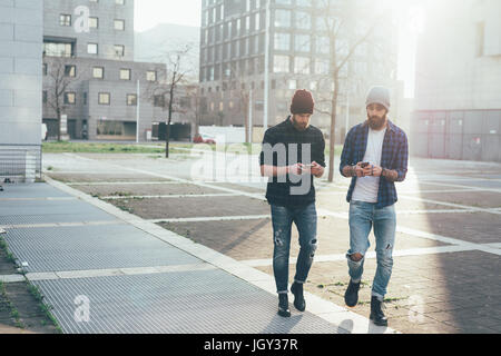 Two young male hipsters walking in city while looking at smartphones Stock Photo