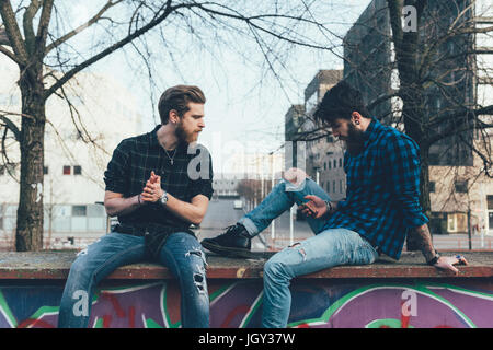 Two young male hipsters sitting on wall looking at smartphone Stock Photo
