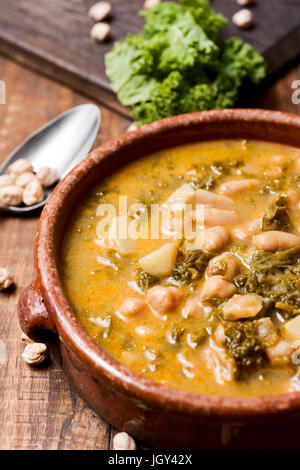 closeup of an earthenware bowl with kale stew with potatoes and chickpeas, on a rustic wooden table sprinkled with some dry chickpeas Stock Photo