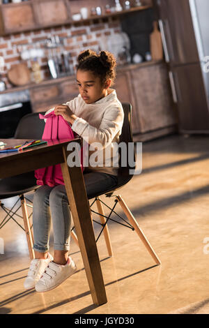 Cute little girl holding backpack while sitting at table with felt tip pens, doing homework concept Stock Photo