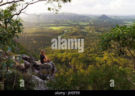 A young man sits, quietly meditating on top of a mountain with expansive views near Noosa Heads, Australia. Stock Photo