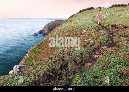A woman in a dress walks on a winding grassy trail beside a cliff above the ocean at sunrise on an isolated coastline near Port Macquarie, Australia. Stock Photo