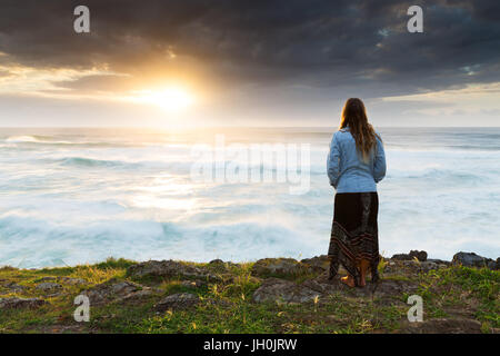 A woman huddles in the cold and watches a bright sunrise over the ocean. Stock Photo