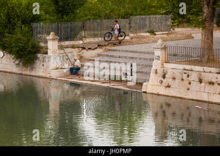 Man fishing of the steps in the grounds of the Royal Palace of Aranjuez in the Madrid Province of Spain watched by boy with bike and a cat Stock Photo