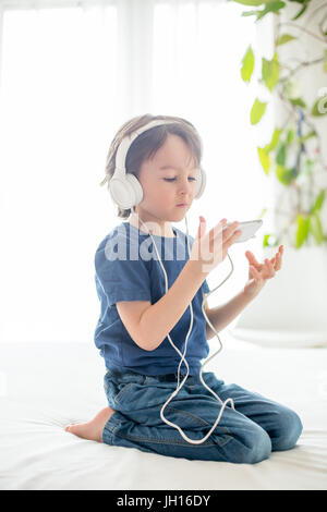 Cute boy with phone and head phones, listening music, indoors, sitting on the bed in bedroom Stock Photo