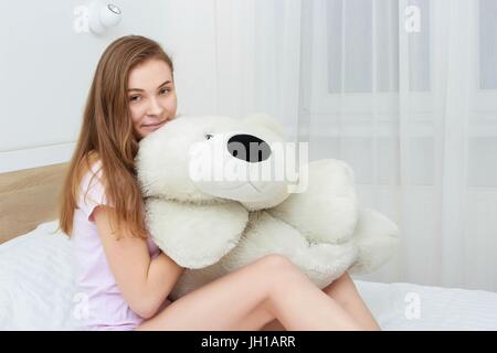 Beautiful teenager hugging soft teddy bear and smiling lying in her bed in the bedroom Stock Photo