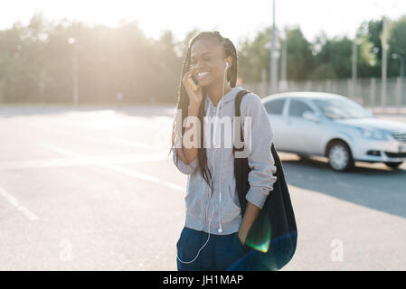 Half-length portrait of the cheerful afro-american teenager talking via the mobile phone while walking along the street during the sunset. Stock Photo