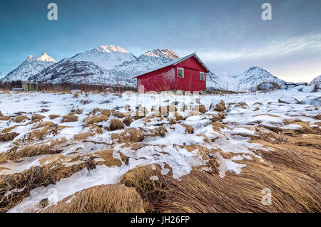 Wood hut framed by rocks covered with grass and ice with snowy peaks in the background, Svensby, Lyngen Alps, Troms, Norway Stock Photo