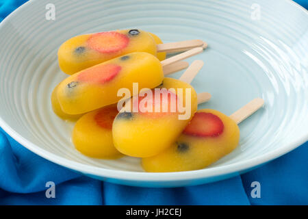 Homemade fruit ice pop, popsicle, ice cream lolly with orange juice, fresh strawberries and blueberries in a  blue porcelain bowl on dark blue fabric  Stock Photo