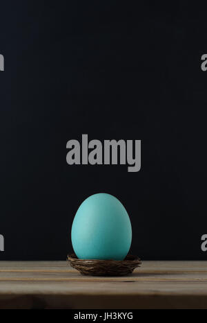 An upright chicken's egg, tinted turquoise blue in small nest on wood plank table.  Black chalkboard background provides copy space above. Stock Photo