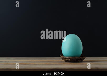 An upright chicken's egg, tinted turquoise blue in small nest on wood plank table.  Black chalkboard background provides copy space. Stock Photo