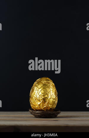 Easter concept.  A chocolate egg, wrapped in crinkled metallic gold foil.  Presented in a small nest on a wood plank table with black chalkboard backg Stock Photo