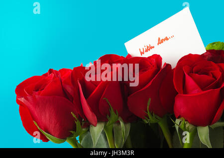 A bouquet of red roses against sky blue background.  A white message card shows 'With Love' in red handwriting. Stock Photo