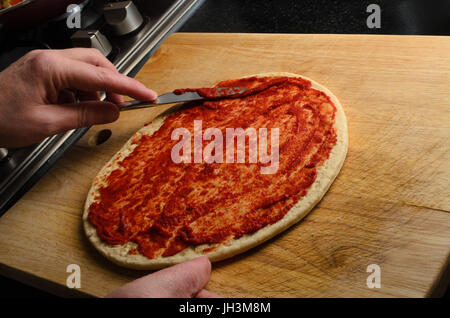 Male hand spreading tomato puree or sauce over pizza base using steel knife on scratched and worn chopping board.  Black work surface and edge of cook Stock Photo