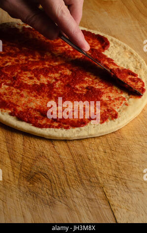 A male hand spreading tomato puree on a pizza base with knife on an old, scratched and worn wooden chopping board.  Dark light. Stock Photo