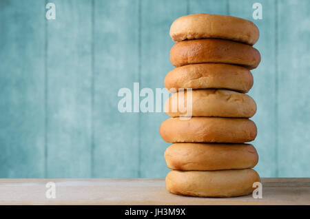 A tall stack of whole bagels, on a light wooden chopping board with a turquoise blue plank background. Stock Photo