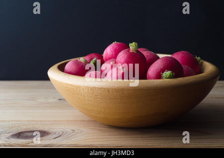 A wooden bowl of  fresh red radishes on plank wood table with black chalkboard background. Stock Photo
