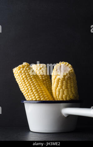 Three sweetcorn cobs, grouped upright in a small white enamel cooking pan on black slate with chalkboard background. Stock Photo