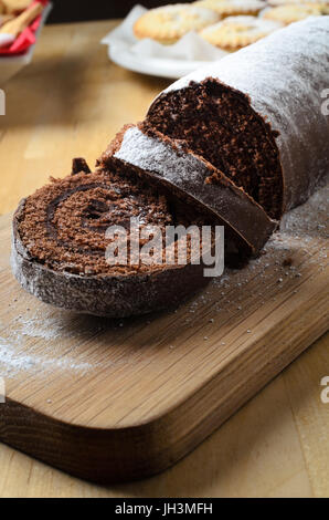 Christmas desserts table with sliced chocolate Yule Log dusted with icing sugar in foreground.  Mince pies and biscuits in background. Stock Photo