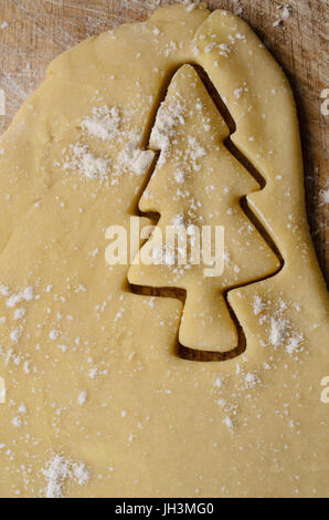 Christmas biscuit or cookie making concept with Xmas tree shape cut out of rolled dough.  Flour sprinkles on pastry and wooden board beneath. Stock Photo